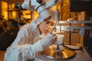 A female researcher in a cleanroom suit uses tweezers to place a material sample under a microscope.