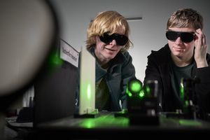 Two boys wearing protective goggles infront of an optical experiment with green lasers. 